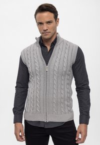 Grau gestrickte Weste mit Kabelmuster, Reißverschluss und geripptem Saum, über einem dunklen Hemd mit Knöpfen getragen, in taillierter Form.