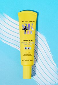 Gul solkremtube merket "Sunray Blur" med SPF 50+, med fargerik tekst og subtile glinsende detaljer, plassert på en blå bakgrunn.