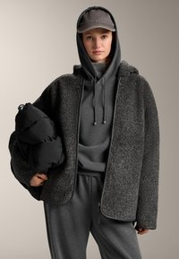 Jachetă gri Sherpa cu fermoar frontal, două buzunare și glugă, purtată peste un hanorac gri cu glugă și pantaloni de trening asortați, ținând o geacă de puf neagră.