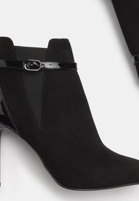 Sorte ankelstøvler lavet af suede, med en glat patenteret læderrem og elastiske paneler, med spids tå og stilettohæle.