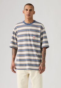 Άνδρας με τατουάζ, φοράει υπερμεγέθη μπεζ και μπλε ριγέ μπλούζα και κρεμ παντελόνι, στέκεται μπροστά σε απλό φωτεινό φόντο.