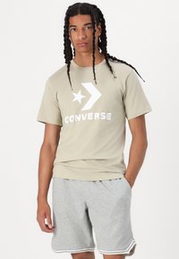 Bežs kokvilnas T-krekls ar lielu baltu "CONVERSE" logotipu un zvaigžņu grafiku, apvienots ar gaiši pelēkiem šortiem ar baltu apdari.