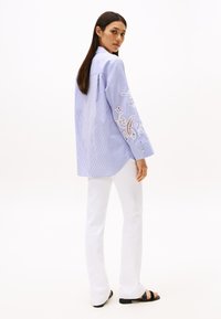 Camicia a maniche lunghe a righe blu e bianche, con dettagli in ricamo bianco su una manica. Abbinata a pantaloni bianchi aderenti e sandali neri.
