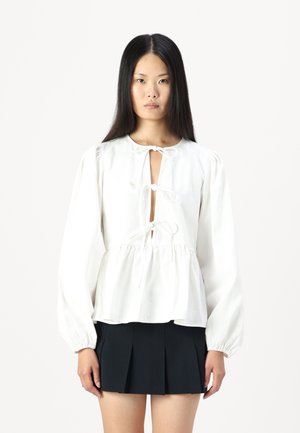 Bluză albă cu mâneci lungi, cu un decolteu în formă de cheie și închideri cu funda, având o talie peplum plisată. Material neted cu un luciu delicat.