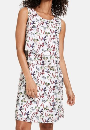 Robe blanche sans manches avec un haut à couches et un imprimé floral comprenant des feuilles et des fleurs violettes, rouges et vertes, portée par une femme aux cheveux bouclés.