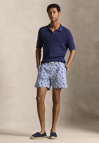 Mladý muž stojí s rukama v kapsách, oblečený v tmavě modrém polo triku s krátkým rukávem, modrých vzorovaných šortkách a tmavě modrých espadrilkách.