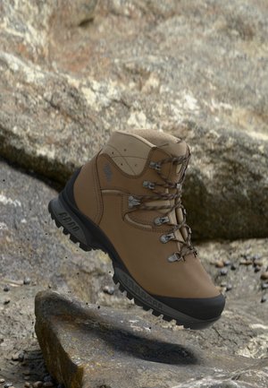Brown hiking boot with black sole and laces, suspended above rocky terrain, showing rugged outdoor design and grip features.