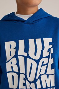 Royal modra kapuca iz mehkega blaga z belim grafičnim tiskom različnih stilov pisave, ki bere "BLUE RIDGE DENIM." Ima kapuco z vrvico.