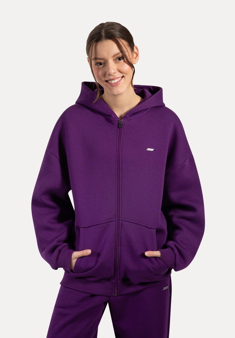 Sudadera con cremallera de color púrpura, hecha de tela suave, con una gran capucha, bolsillos frontales y puños acanalados. Logo de la marca visible en el pecho.