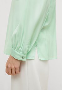 Blusa di seta verde menta con maniche lunghe. Presenta polsini arricciati e spacchi laterali all'orlo, accompagnata da un bottone metallico sul polsino.