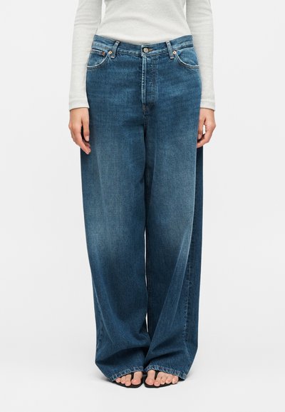 Personne portant un jean denim bleu à jambes larges, associé à un haut blanc à manches longues, debout pieds nus sur un fond uni.