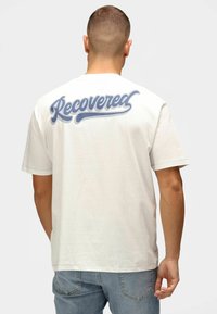 Hvid bomuld t-shirt med et stort blåt kursiv "Recovered" grafik på bagsiden. Korte ærmer, rund hals, afslappet pasform.
