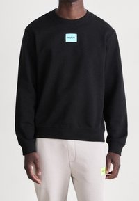 Sudadera negra de algodón con cuello redondo, puños acanalados y una etiqueta rectangular azul con "HUGO" en el frente.