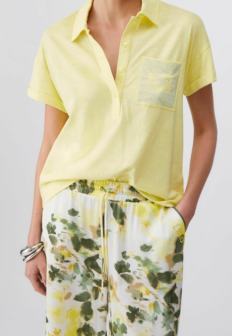 Personne portant une chemise jaune à manches courtes avec poche poitrine et un pantalon blanc à motif floral vert et jaune avec taille à cordon.