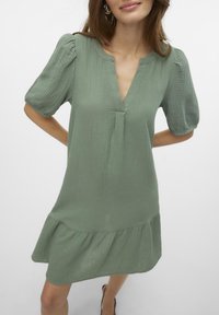 Svijetlozelena haljina od teksturiranog materijala, s V-izrezom, kratkim napućenim rukavima i volanima na rubu koji su slojeviti.
