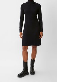 Robe noir à manches longues à côtes, avec un col montant, présentant une silhouette droite et une longueur genoux, associée à des bottes noires épaisses à la cheville.