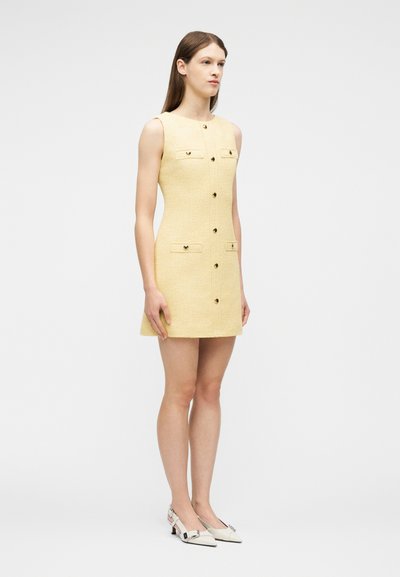 Γυναίκα που φοράει μίνι φόρεμα χωρίς μανίκια σε κίτρινο χρώμα με τέσσερις τσέπες με καπάκι και κουμπιά μπροστά, σε συνδυασμό με λευκά μυτερά παπούτσια με χαμηλό τακούνι, όρθια.