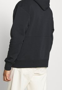 Felpa con cappuccio nera realizzata in morbido tessuto, caratterizzata da una vestibilità comoda, polsini a coste e un dettaglio di cucitura lungo la schiena.