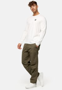 Weißes Langarmhemd mit kleinem schwarzem Logo auf der Brust, kombiniert mit olivgrünen, lässigen Cargo-Hosen und weißen Sneakers mit Reißverschlüssen.