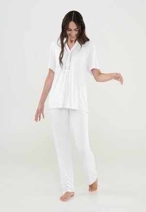 Mujer con cabello largo y oscuro sonriendo, vestida con un top blanco de manga corta y pantalones a juego, caminando descalza sobre un fondo claro y sencillo.