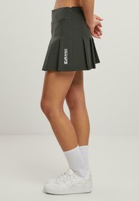 Grauer, gefälster Rock mit einem Taillenbund und einem Reißverschluss. Mit weißem Branding an der Seite. Kombiniert mit weißen Sneakern und Knöchelsocken.