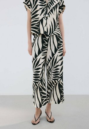 Frau trägt lockere schwarz-weiße, blattgemusterte Cropped-Hosen und ein passendes kurzärmeliges Oberteil mit schwarzen Riemchensandalen.