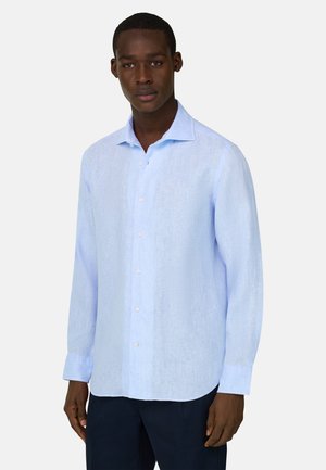 Nuori mies, jolla on vaaleansininen pitkähihainen nappipaita ja tummat housut, seisomassa yksinkertaisen vaalean taustan edessä.