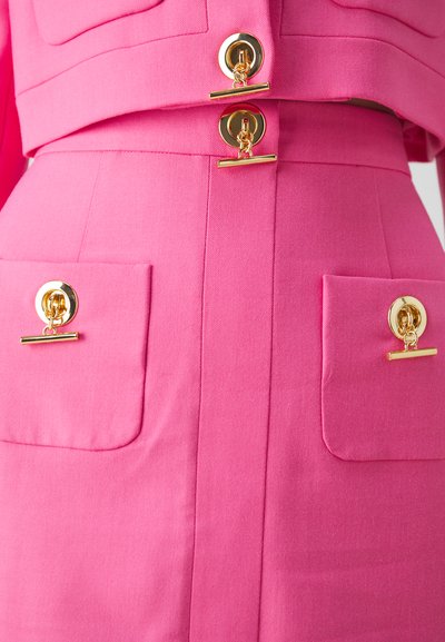 Veste rose sur mesure avec jupe assortie. Comprend des boutons en métal doré et des poches avec des accents en chaîne. Tissu à texture lisse.