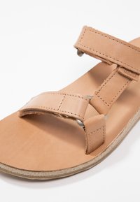 Sandale en cuir couleur fauve avec des brides cousues et des rivets métalliques sur une semelle claire, vue en gros plan sur fond blanc.