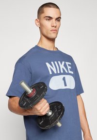 Blå kortærmet t-shirt med stort hvidt "NIKE" logo og nummer "1." Holder en sort vægt med metallisk håndtag og røde accenter.
