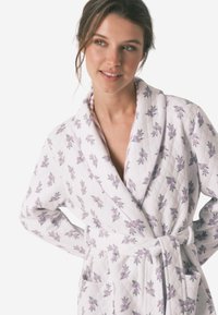 Accappatoio bianco con motivo floreale, realizzato in tessuto trapuntato. Presenta un colletto a scialle, una cintura in vita e tasche, con decorazioni di fiori viola e verdi.