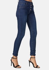 Pantalones ajustados en denim azul oscuro con cinco bolsillos, cierre de botón y costuras en contraste. Combinados con tacones altos negros con tiras.