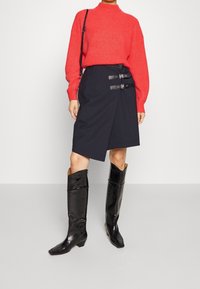 Vrouw in een rode gebreide trui, zwarte asymmetrische rok met dubbele gespen, zwarte kniehoge laarzen en een zwarte schoudertas.