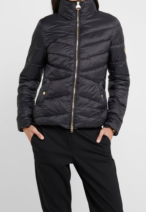 Woman wearing a black quilted jacket with gold zipper and button details, hands in pockets of black pants against a plain light background.