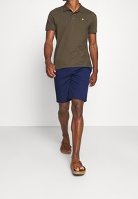 Polo vert olive avec col, shorts navy et sandales marron. Le polo arbore un petit logo jaune sur la poitrine.