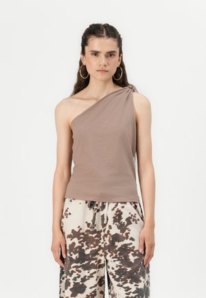 Brown one-shoulder top made of soft fabric with a twisted accent. Paired with cream pants featuring a brown cow print pattern.