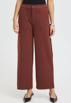 Femme portant un pantalon marron taille haute à jambes larges, des talons noirs à bouts pointus et un haut noir, debout avec les mains détendues le long du corps.
