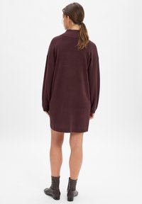 Sukienka dzianinowa w głębokim odcieniu bordowym, z wysokim kołnierzem, długimi rękawami i luźnym krojem. Długość do połowy uda, zestawiona z brązowymi botkami za kostkę.