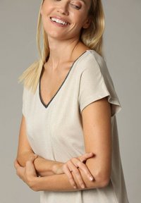 Haut à manches courtes, beige, avec un décolleté en V, arborant une légère brillance et un bord sombre contrastant le long du décolleté. Texture douce.