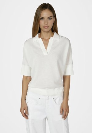 Woman with straight hair wearing a white short-sleeve collared top and white pants, standing with arms relaxed against a plain background.