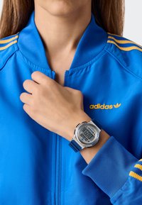 Ezüst színű tokú digitális óra, kék szilikon szíjjal és digitális kijelzővel, amely az időt mutatja. Egy kézen viselik, amelyen egy kék cipzáras dzseki van.