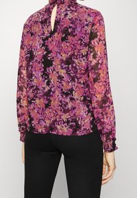 Blusa trasparente floreale con un design nero, viola e arancione; presenta un collo alto, polsini arricciati e un'apertura a chiave sul retro.