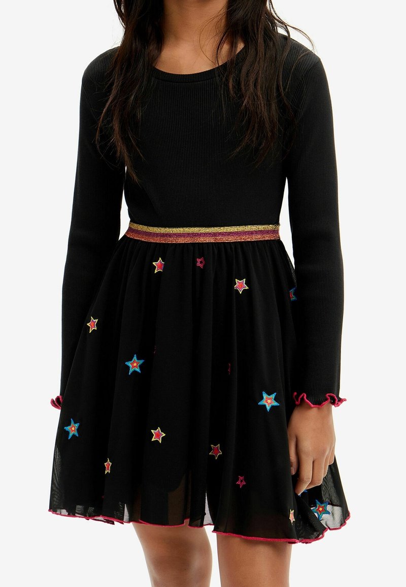 Fille portant une robe noire à manches longues avec des étoiles brodées colorées et une ceinture rayée scintillante.