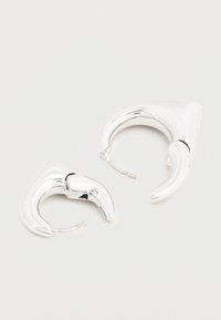 Kaks säravat hõbedast rõngaskõrvarõngast keeruka, skulpturaalse disainiga tavalise valge tausta ees.