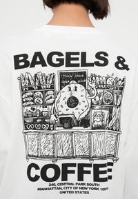 Κάρικας ντόνατ πίσω από πάγκο αρτοποιείου με ψωμιά, πρέτζελ και κούπες καφέ, με έντονο κείμενο «Μπέιγκελς & Καφές» και διεύθυνση στη Νέα Υόρκη από κάτω.