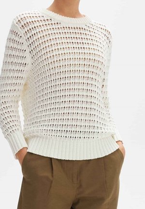 Personne portant un pull blanc en maille ajourée avec des poignets et un ourlet côtelés, associé à un pantalon marron taille haute, mains dans les poches.