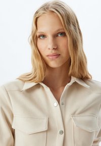 Camisa de botón color beige claro hecha de un tejido suave. Cuenta con dos bolsillos en el pecho, botones de metal plateado y un cuello. Textura suave y diseño clásico.