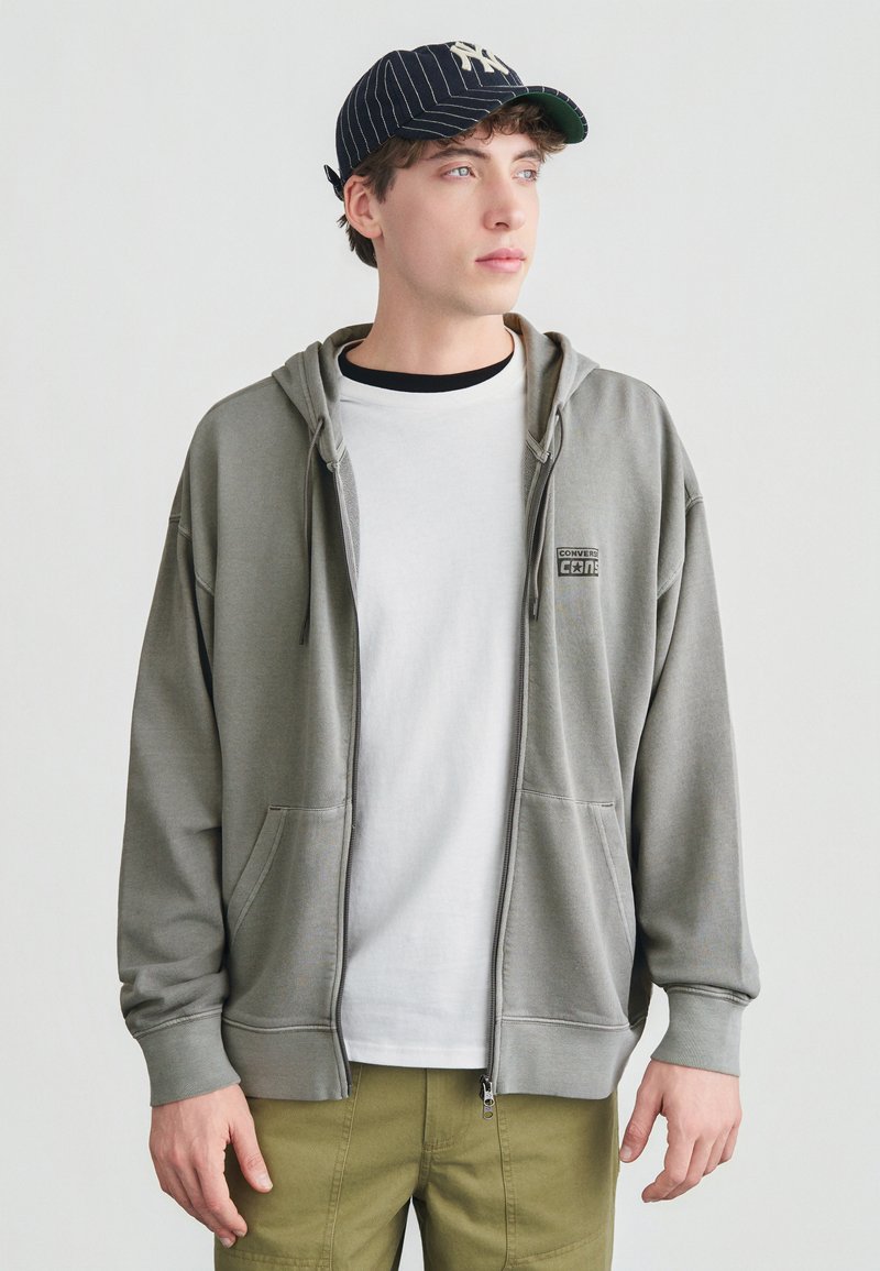 Νεαρός άνδρας που φοράει ριγέ καπέλο μπέιζμπολ, γκρι ανοιχτό φούτερ με φερμουάρ και μπροστινές τσέπες, λευκό T-shirt και χακί παντελόνι, κοιτάζει δεξιά.