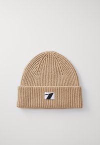 Beige gebreide muts met een geribbelde textuur, voorzien van een omgevouwen rand en een klein zwart-wit logo op de voorkant.