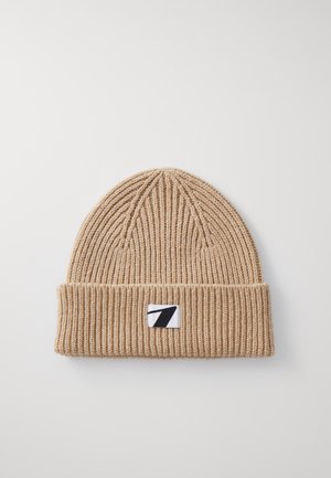 Bonnet en tricot beige avec une texture côtelée, comportant un revers et un petit patch logo noir et blanc à l'avant.